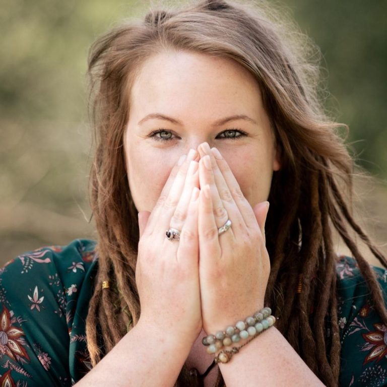 Junge Frau mit Dreadlocks, die lächelt und die Hände vor dem Gesicht hält.