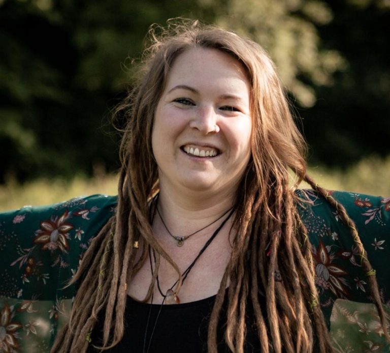 Lächelnde Frau mit Dreads, stehend in einer grünen Umgebung, trägt ein Blumenshirt.