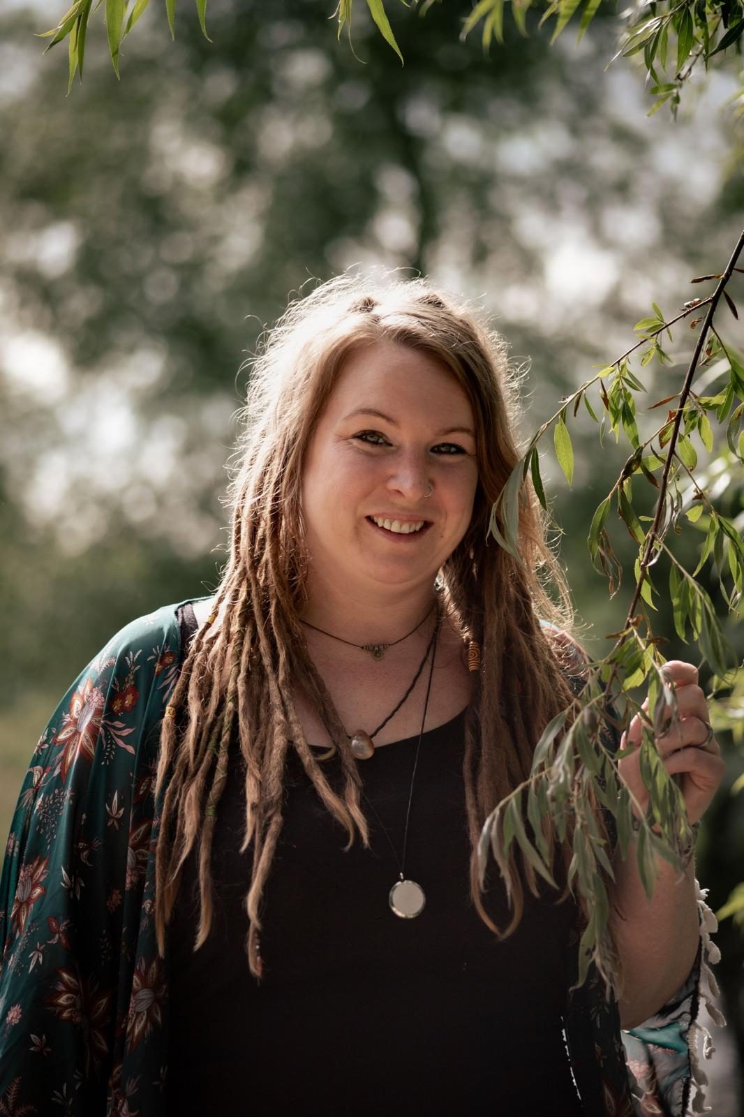 Lächelnde Person mit Dreadlocks, die an einem Baum steht und eine Blätterzweig hält.